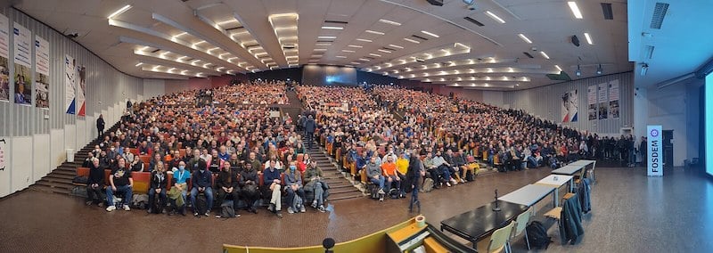 Alt: A huge room where keynote takes place is full of hundreds of people sitting in theatre style sirring arrangement. There is sign at the right end of the picture with FOSDEM written in big purple letters.