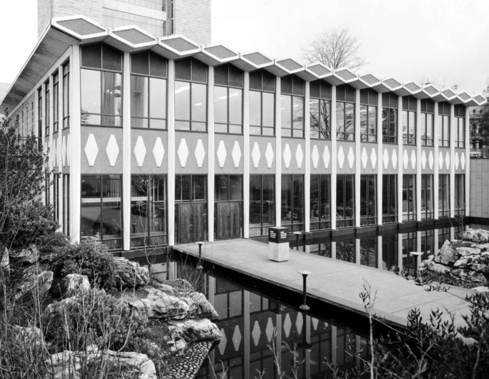 Black and white photograph of the provincial archives building. It has two stories and glass windows in the front. There is a causway over water that leads to the entrance.