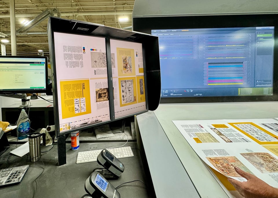 Two displays at a printing company showing, left, a preview of the sheet being printed using digital files, and, right, ink levels and other pres controls. A hand at lower-right lifts up a printed sheet being examined for quality.
