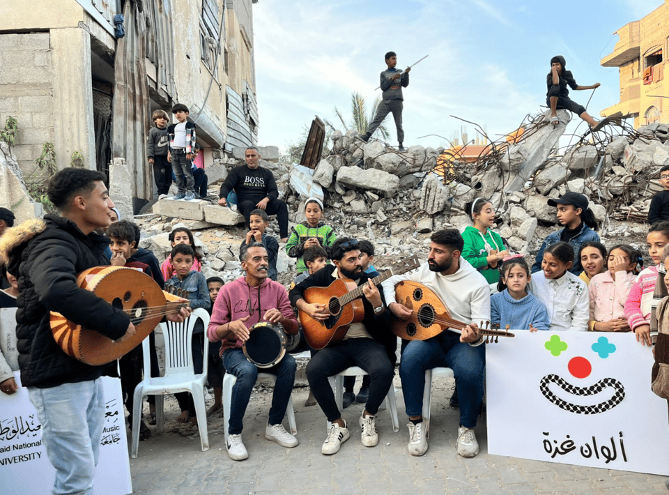 Four people playing musical instruments, with children nearby, in front of rubble