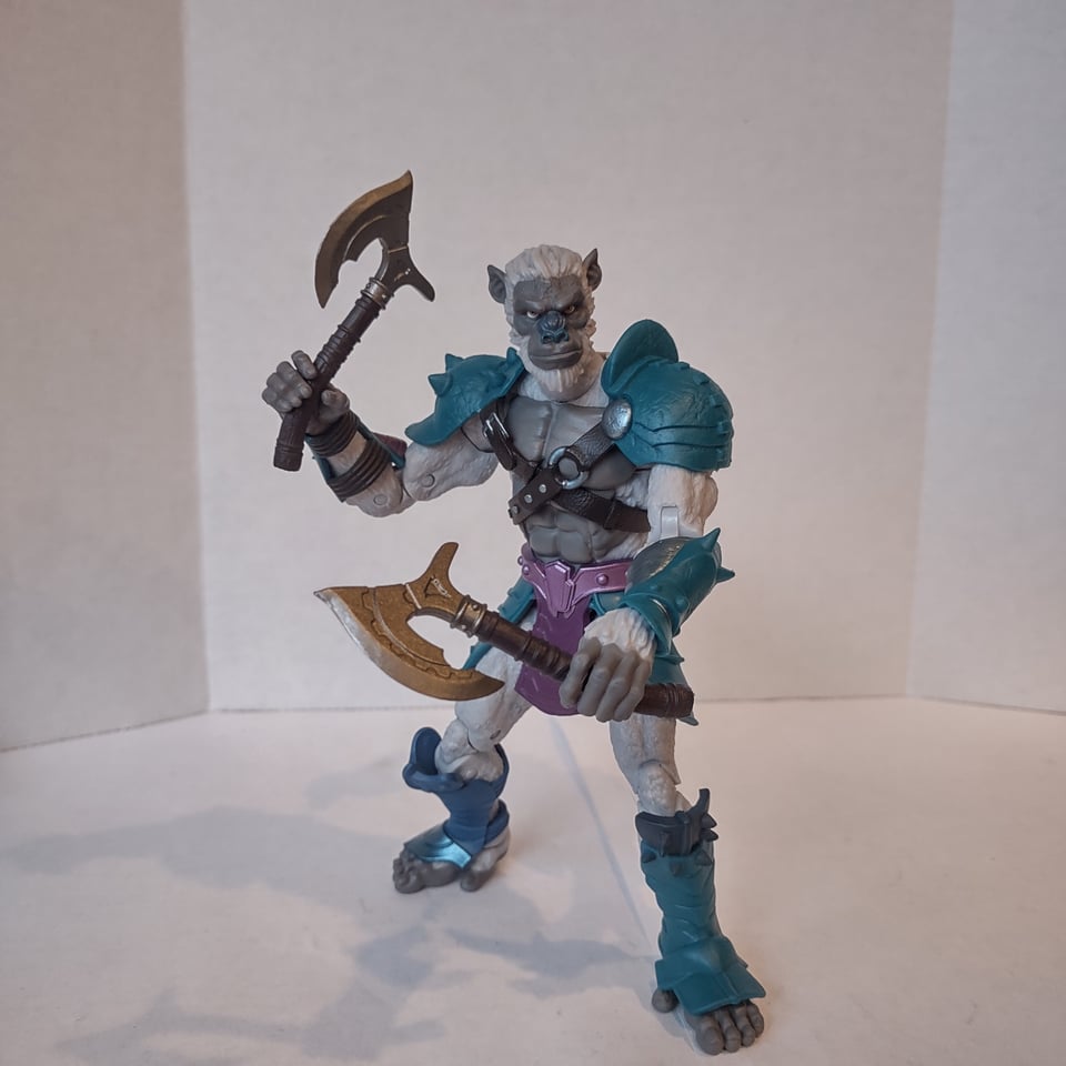 An action figure of a white -furred ape-man. He wears sections of blue and purple, fantasy-styled armor and holds an ornate axe in each hand