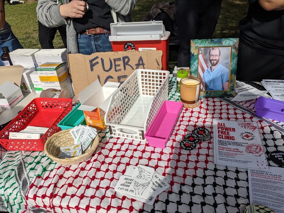 A table covered with a black, red, green and white keffiyeh and scattered with flyers and stickers for the All Power Free Clinic. A cardboard sign reads FUERA ICE. There is a candle next to a framed photo of Alex Pretti. There are red and white baskets of medical supplies.