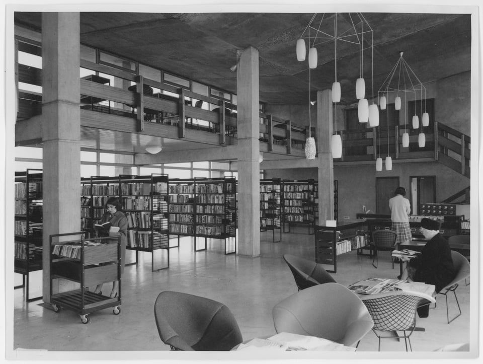 An internal photo of the original building from the north west corner. Light is coming in from the eastern curtain wall to illuminate metal bookshelves. H shaped beams hold up the student gallery. Oval frosted glass lights hang over an issuing desk, and wire-frame metal armchairs are set around tables.