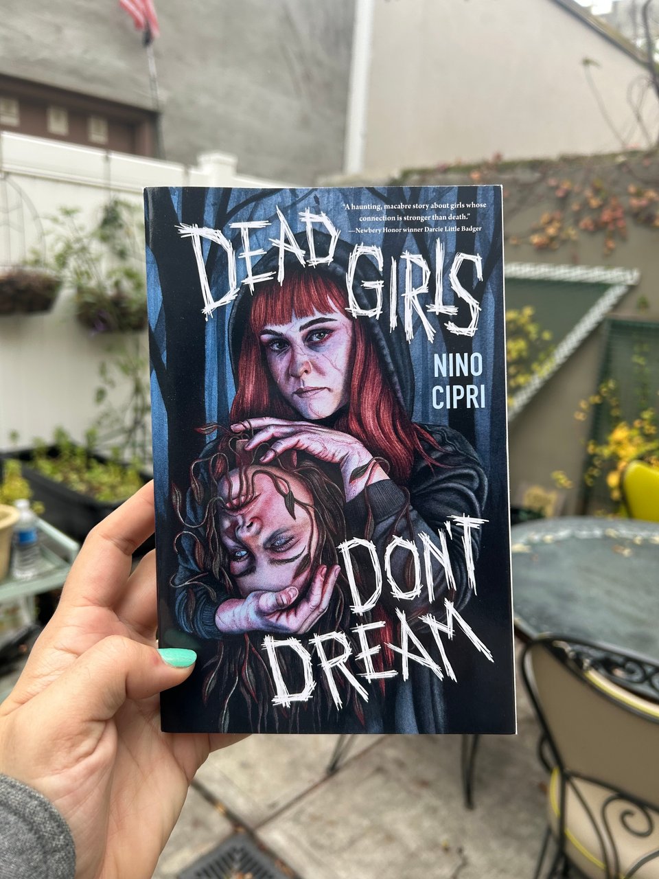 Nino's hand holding their book Dead Girls Don't Dream in a garden patio.