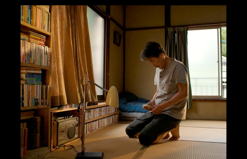 A man kneeling on the a tatami floor in a Japanese apartment. He's looking at a book and kneeling next to a shelf of books, cassettes and a small cassette player.