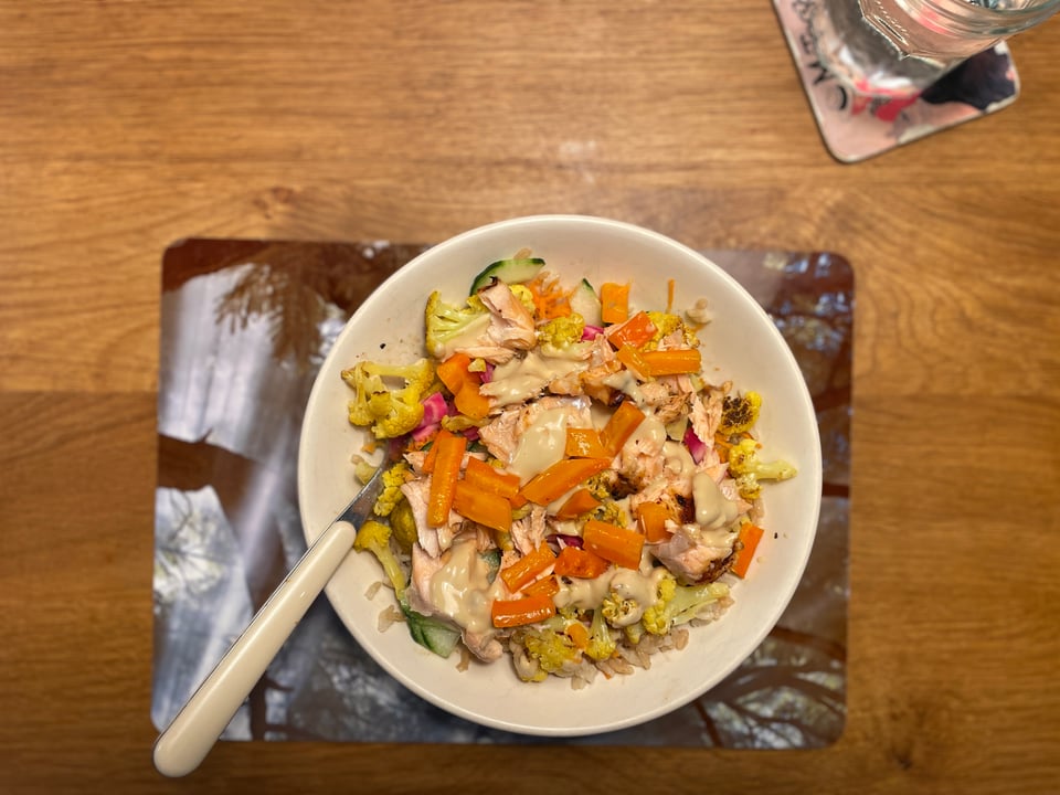 A bowl full of roasted veggies and cooked salmon sits on a placemat on a wooden table.