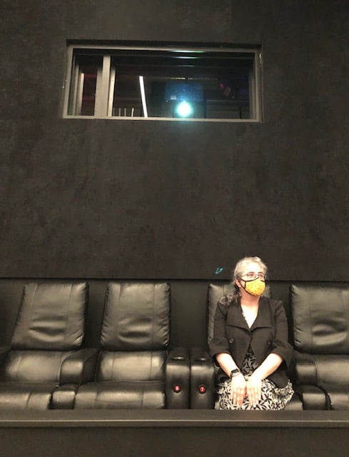 A photo of your intrepid newsletter-writer, masked, alone in a movie theater, looking pensively at something off camera, with the projector in shot above and behind her