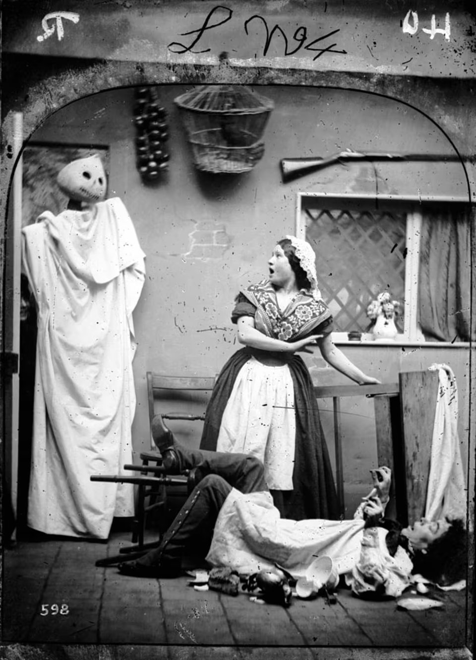A vintage staged photo showing two people acting terrified by a figure in the corner with a pumpkin-like head, draped in a sheet. A woman turns, startled, and a man lays on his back as if his chair has been knocked over