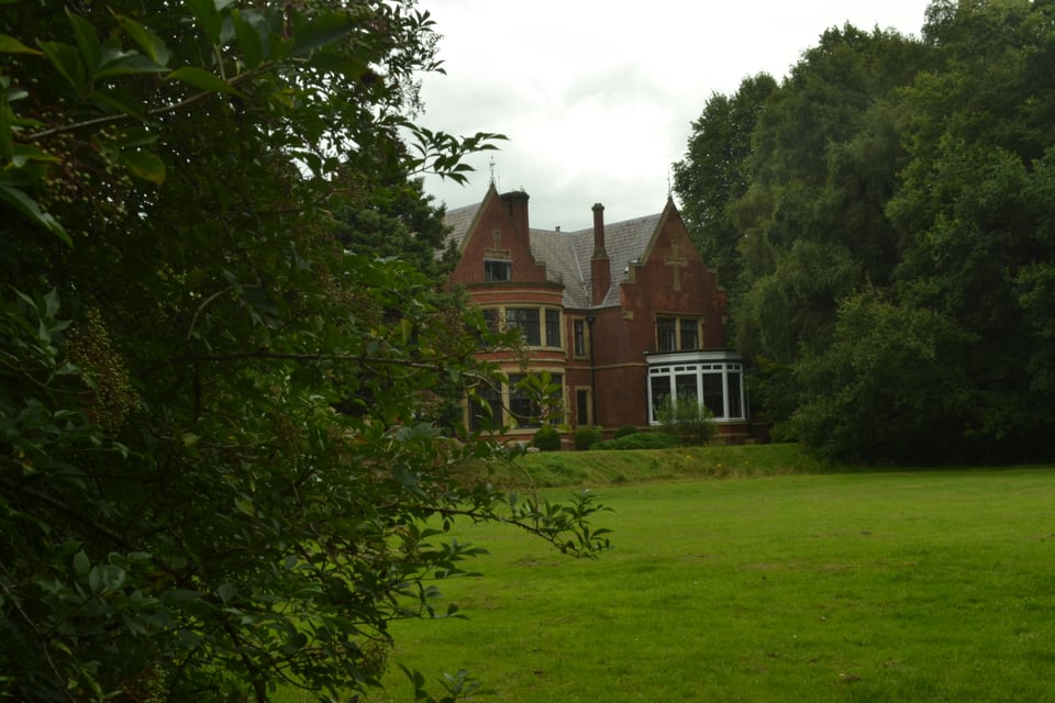 A red brick mansion with large bay windows peeks through trees. In front is a green expanse of lawn.