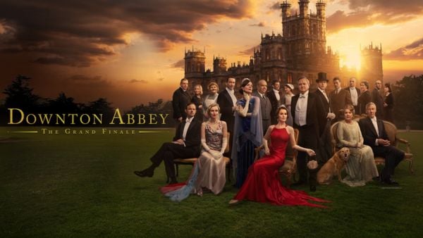 Photo: Promotional image for Downton Abbey: The Grand Finale. The cast, dressed in formal evening wear from the 1920s, are arranged on a manicured lawn in front of Highclere Castle at sunset. The castle's distinctive towers are silhouetted against dramatic orange and grey clouds. In the foreground, a woman in a striking red gown reclines on the grass, while other cast members stand and sit in elegant poses wearing period dress in various colours including beige, blue, and cream. The title "Downton Abbey: The Grand Finale" appears in gold lettering at the bottom left.