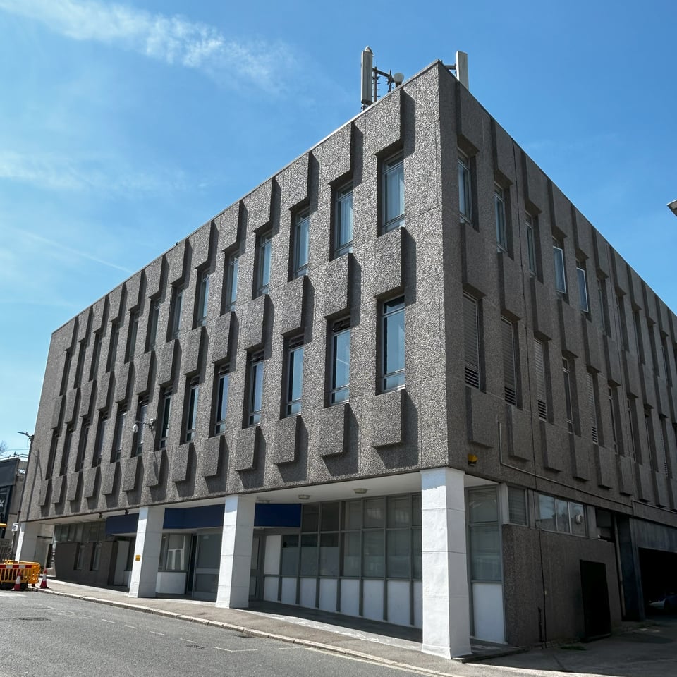 A brutalist office block. The rough concrete walls have regularly set out windows with blocks jutting out to form bas reliefs. It is intensely regular. At the ground level, the glass wall has been set back and a rather random doorway added.