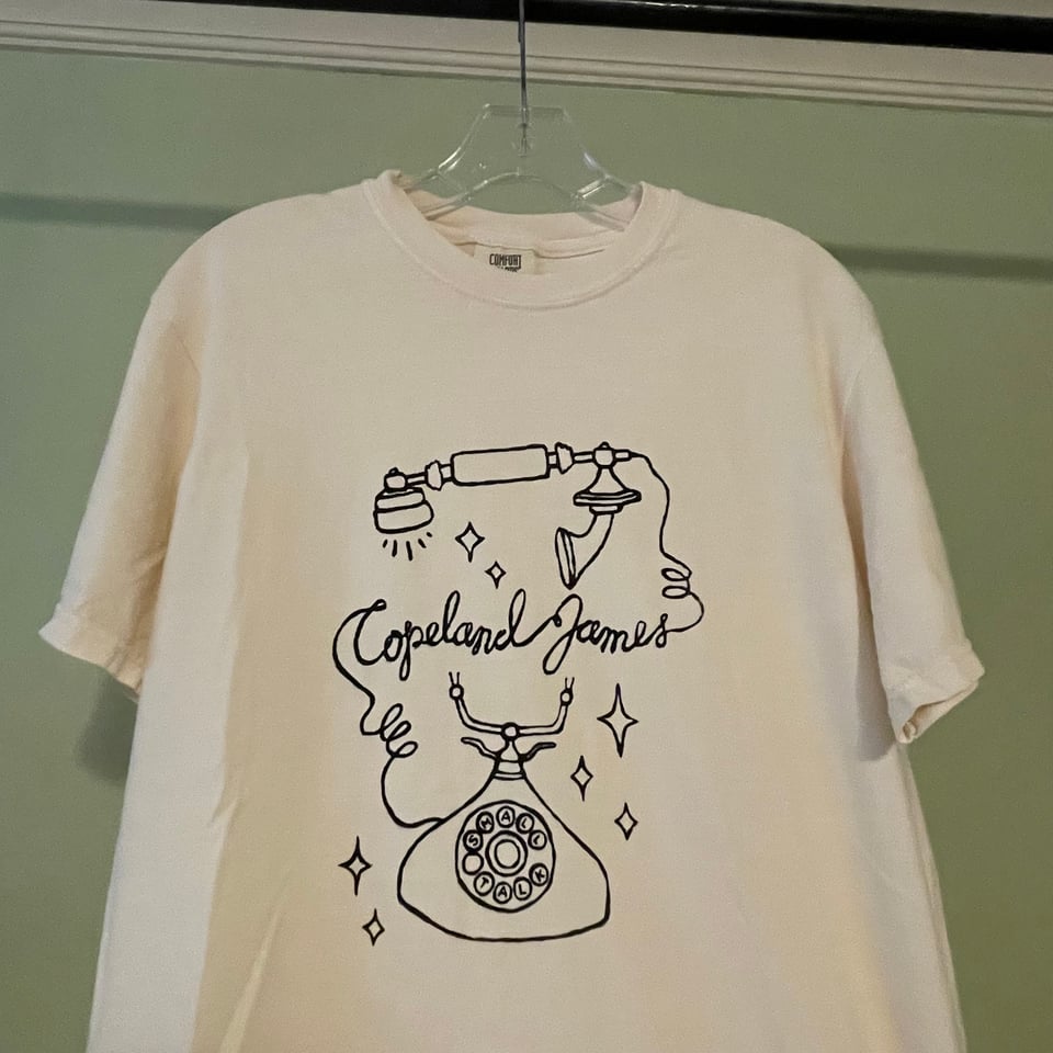 A Copeland James T-Shirt is displayed on a hanger. The shirt is cream-colored, with a vintage rotary phone screen-printed on it in black ink. The receiver is floating above the base of the phone and beneath it, the cord curved to spell "Copeland James" in cursive. Instead of numbers, the dial has letters that spell out "Small Talk."