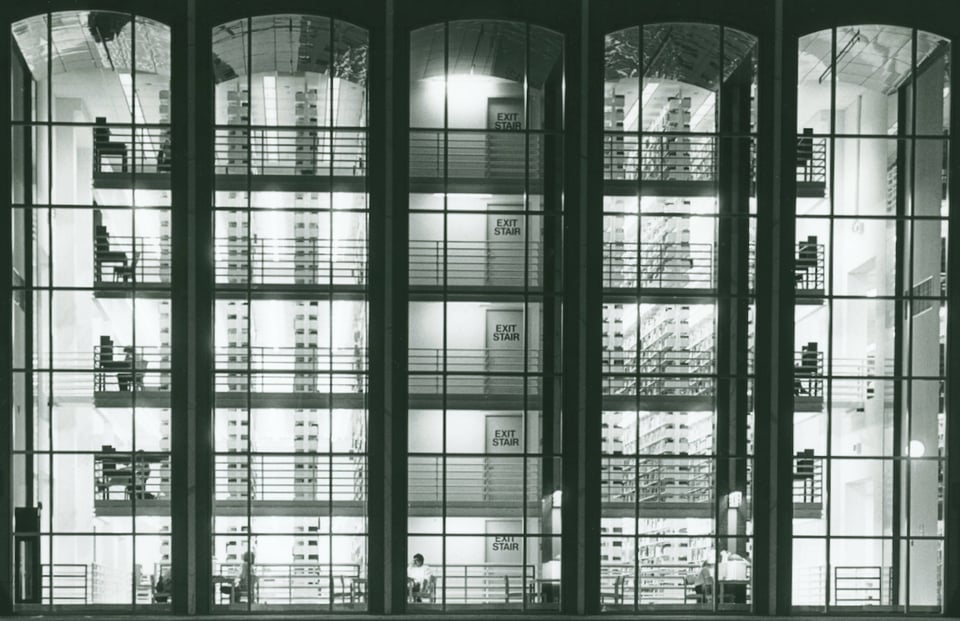 A black and white photograph of a library at night from the outside, with large glass windows showing the stacks and people studying