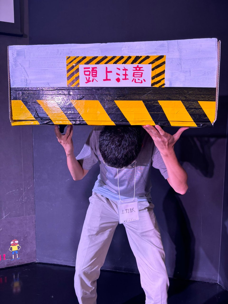 A person holds up a cardboard box painted to look like a low, overhanging ceiling, with a warning sign and yellow and black striped caution tape. The person crouches underneath it as if they can't stand up fully.