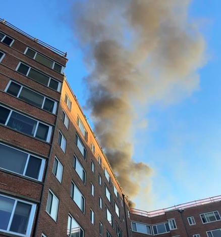 A brick apartment building with smoke billowing from the roof against a blue sky