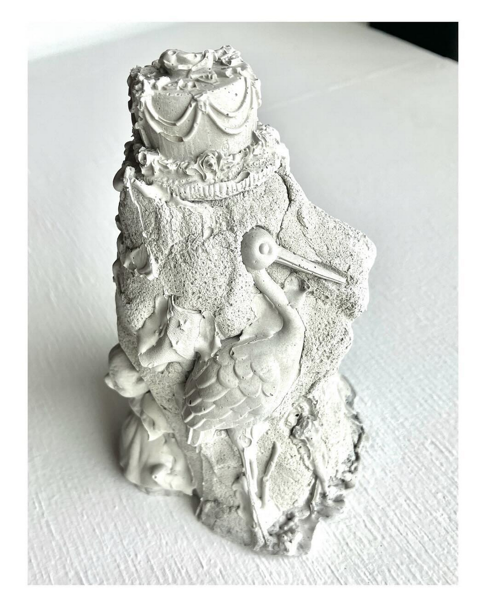 a roughly cast concrete sculpture featuring the likeness of a cartoon stork and a birthday cake, among other objects