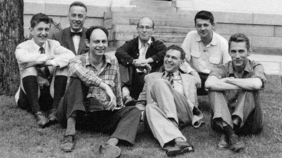 A black and white photo of seve white men in their 20s to 40s sitting on a lawn in front of a building.