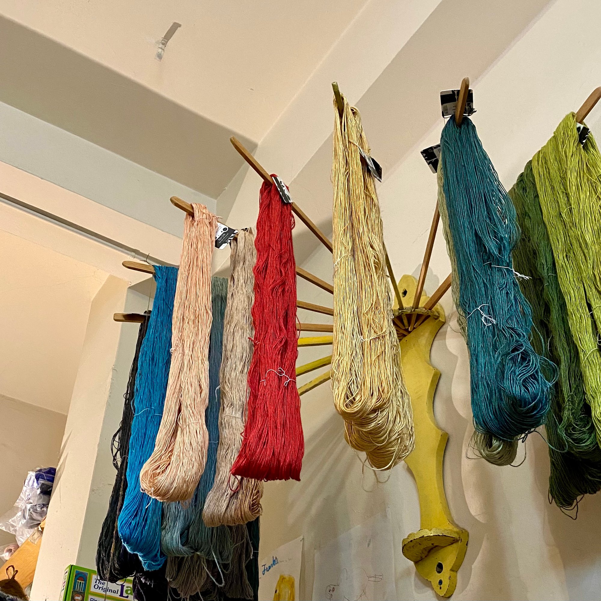 Image of deep blue, peachy pink, red, wheaty yellow, and kelpy green yarn hanging on a yellow rack.