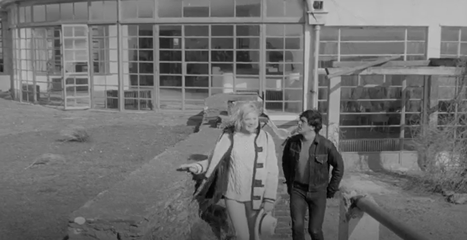 screenshot with two actors walking up some steps. Behind them is the glass wall of the palm court, with a plain wall of Crittall windows in a simple grid. the whole place looks abandoned.