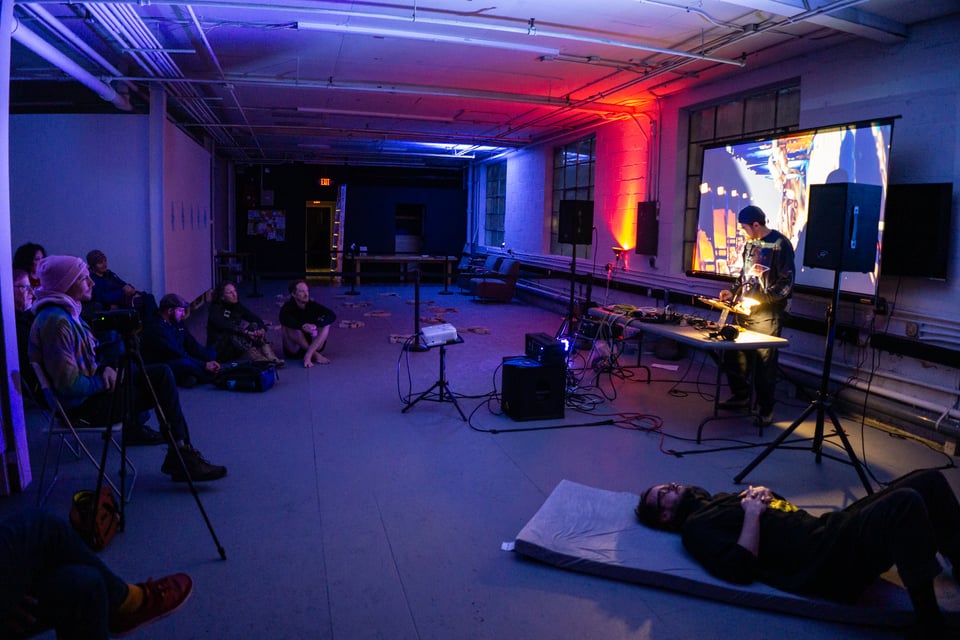 An art gallery with dim, colorful lighting, a projector screen, PA, and people enjoying live music.