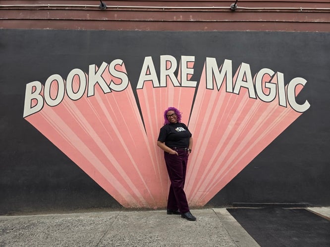Patricia in purple trousers and a black shirt that has Peppermint Patty on it and says the word, “sir.” Patricia is standing in front of a large mural that says “Books Are Magic.”