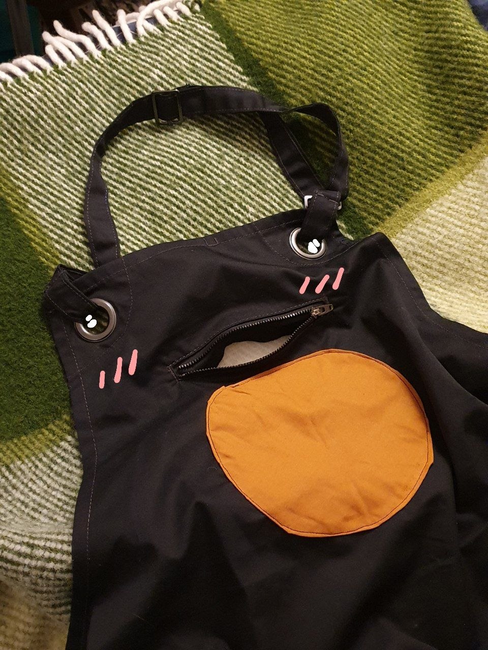 The chest of the apron showing the orange circle, loose threads now cleaned away. The open pocket above the circle looks like a mouth, and the metal circles to which the neck strap is attached look like eyes. I've drawn blushing cheeks beneath the eyes to make the apron make a demure face expression.