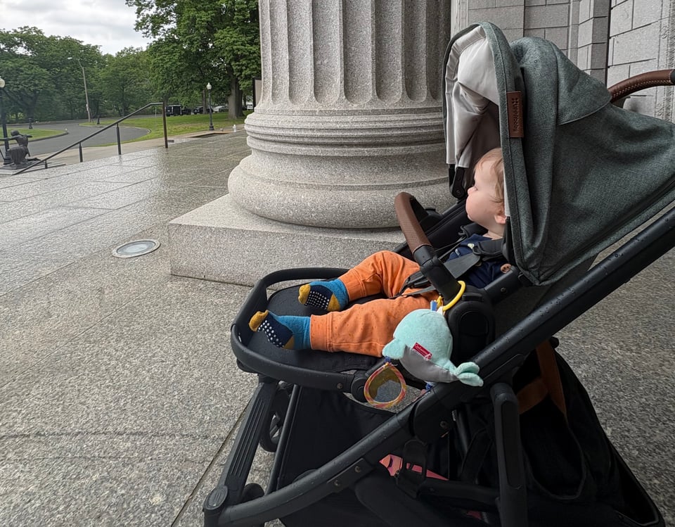 Baby in a stroller beneath the roof of the MFA, can see it's raining just past the columns