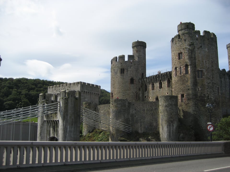 The walls and towers of a castle, with two big round towers with turrets. In the foreground is a suspension bridge anchored to the castle, and the railing of a road bridge.