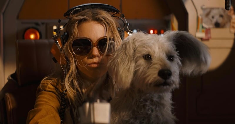 A young woman wearing large sunglasses and headphones sits in a cockpit-like setting with a small dog beside her.