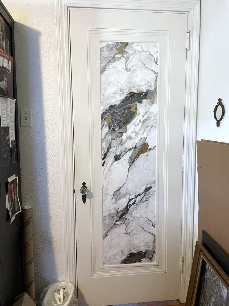 A white door with a panel in the center with white, black, grey, and gold marble patterning on it. To the left is a black piece of furniture with various papers taped to it. To the right, on the floor art and frames are stacked against the wall.