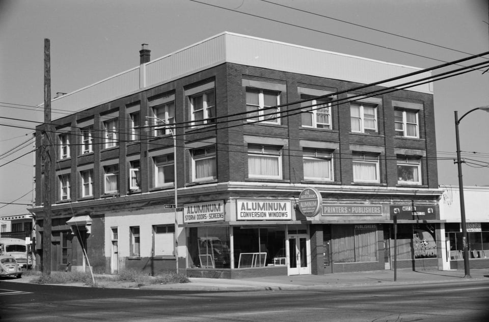 Black and white image of a corner lot with a three story building. A sign saying "Aluminum Conversion Windows" is most prominent.