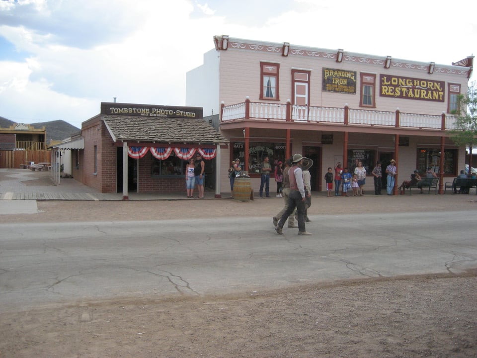 A dusty desert street. On the far side is a two storey building labeled "Longhorn Restaurant" and a small shack labeled "Tombstone Photo Studio." Two men dressed as cowboys are walking down the middle of the street.