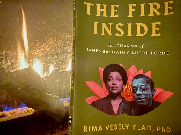 Closeup of a green book cover next to blue and red flames in a gas fireplace. The book cover has yellow and white text and the faces of Black writers Audre Lorde and James Baldwin on top of a pink lotus flower. The text reads “The Fire Inside” “The Dharma of James Baldwin & Audre Lorde” “Rima Vesely-Flad, PhD.”