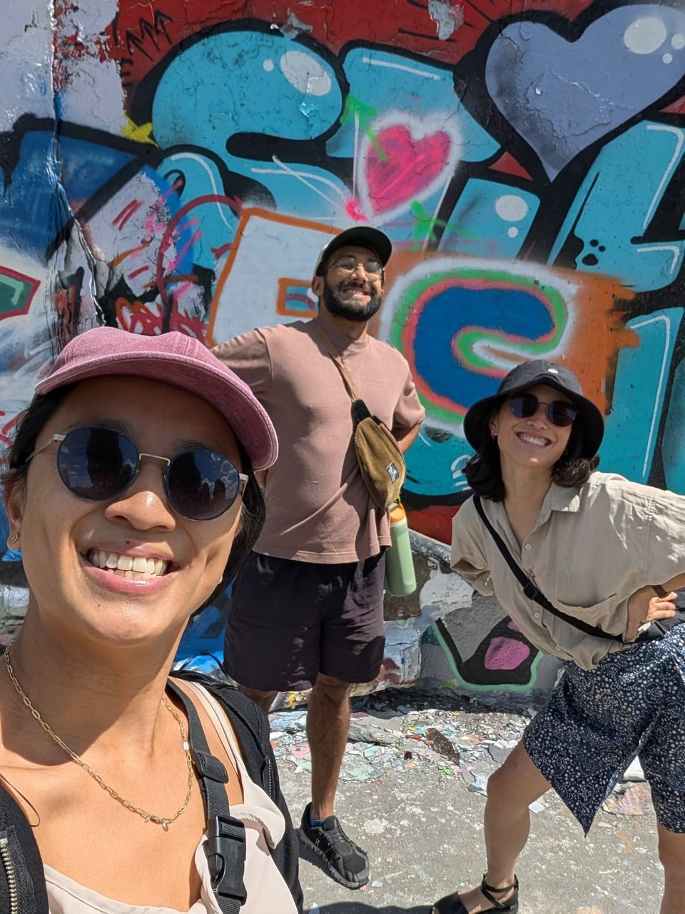 Three people in sunglasse and summer clothes smiling in front of a heavily graffitied wall.