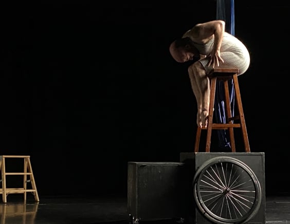 In a black void, I am on top of a wooden stool on top of a large black wooden box. I'm sitting sideways, curled over my legs, arms gripping my thighs with splayed fingers, toes tightly curled below. There is a wheelchair wheel against the box, a smaller box beside, an a distant short stepladder. Everything is starkly lit from the side.