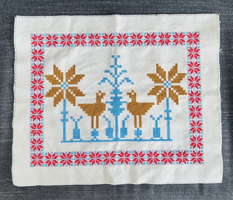 A piece of stitchwork on a white cloth, with scene showing two yellow birds facing each other, surrounded by blue plants and yellow flowers. The scene is surrounded by a square stitchwork frame consisting of red flowers on a blue background