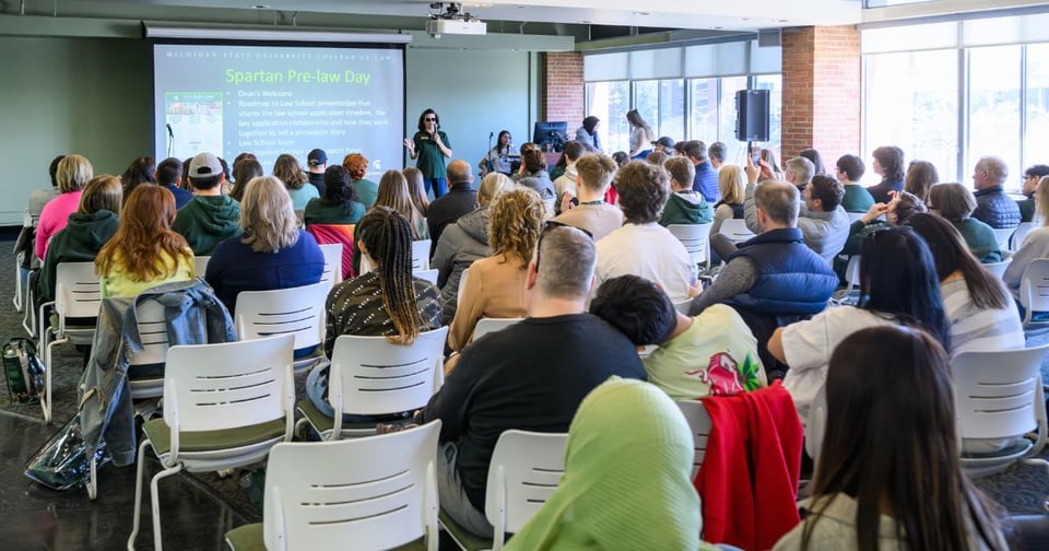Speaker with a microphone, presenting in front of a screen that reads "Spartan Pre-Law Day" to a large crowd of people.