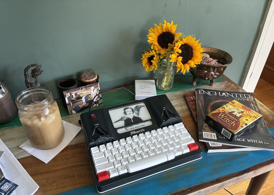 An image of my writing desk; including Freewrite typewriter, sunflowers, magazines, oracle cards, gold cauldron, iced coffee, candles, photo of my grandmother at my grandfather's grave.
