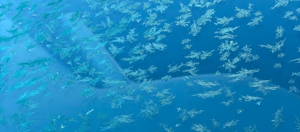A car windshield in various shades of blue, with scattered long frost crystals on it