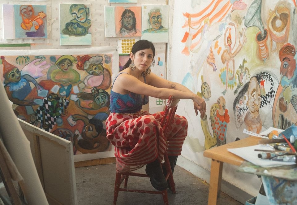 A woman with her long hair pulled back, wearing a blue-and-red patterend tank top and a pink skirt with red polka dots sits facing backward on a chair in the corner of a room with art on the walls.