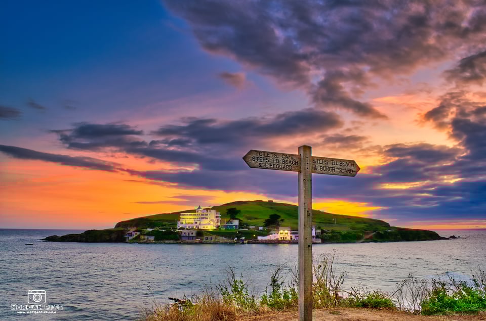 A small island with a purple and orange sunset behind it. On its near shore, a white four storey hotel is light up, glowing amber. In the foreground, on the mainland shore, a finger signpost points towards various locations.