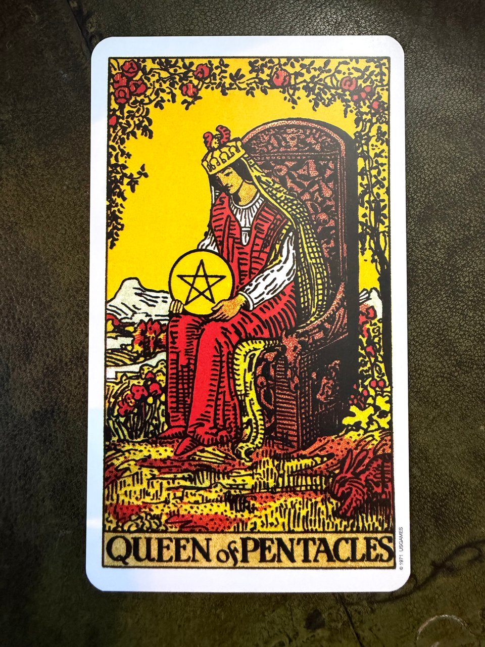 A woman in a red dress, golden crown, and golden veil sits on a wooden throne, tenderly looking down at a golden, pentacle coin in her lap. She's surrounded with lush flowers and greenery.