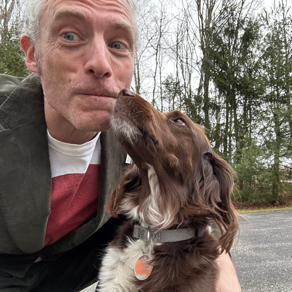Photo of brown and white spaniel/terrier sorta mix dog about to smooch a white man who's wearing a white T-shirt with a red circle, and a green corduroy suit