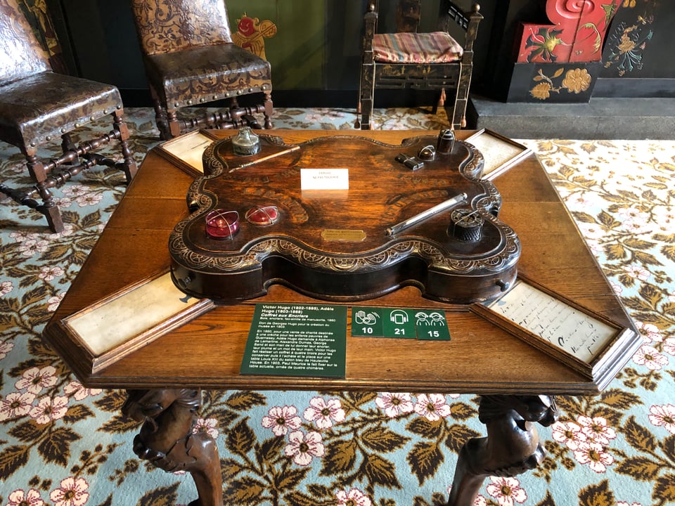 A square-ish wooden table with a low chest with sinuous edges above it. Four inkpots, two quills, and framed four texts appear at the four corners. The table sits on a hideous carpet patterned with flowers and foliage. In the background, medieval style chairs (leather with studs) and a chinoiserie-style screen are partly in the frame.
