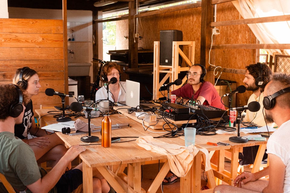 [People sitting around a wooden table in a sun soaked room, wearing headphone and talking into microphones.]