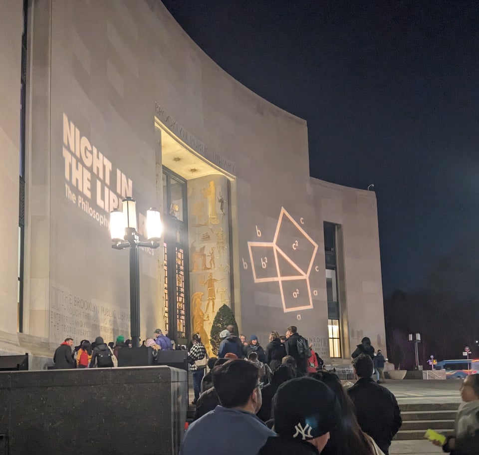 the front of the Brooklyn Public Library Central branch with a ton of people in line out front of it, night just falling. the walls have light-up signage that say "night in the library" along with an image of a right triangle with squares on each of its sides and the pythagorean theorem symbols around it.