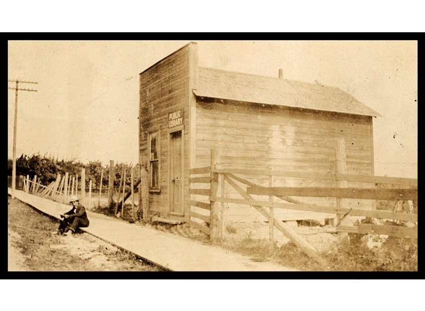sepia toned photo. A person sits on a wooden boardwalk in front of a small wooden building with a sign that reads "Public Library"