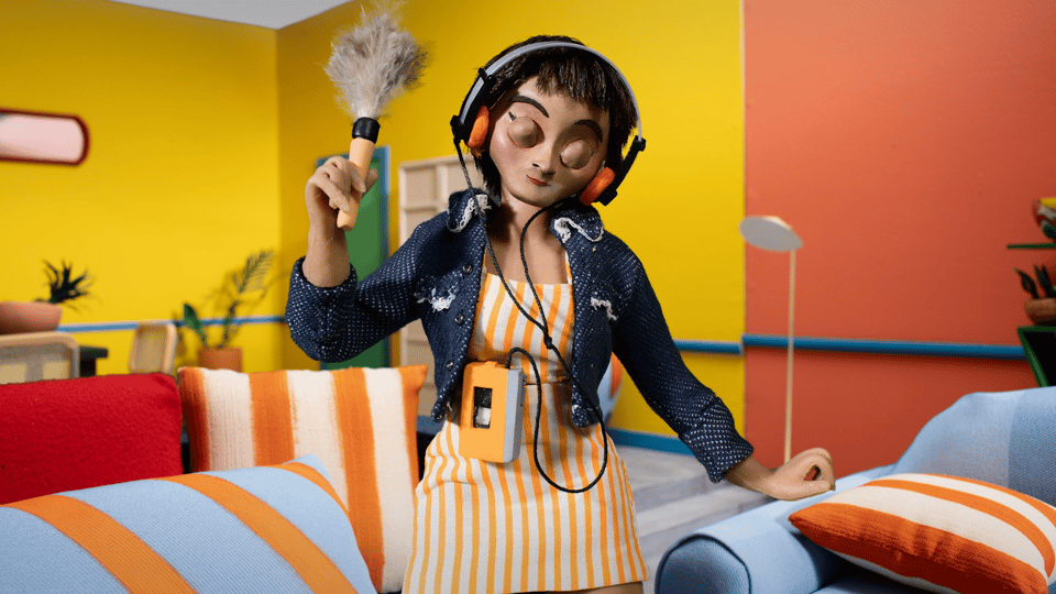 A woman stands in a brightly colored living room, wearing headphones and dancing while holding a duster