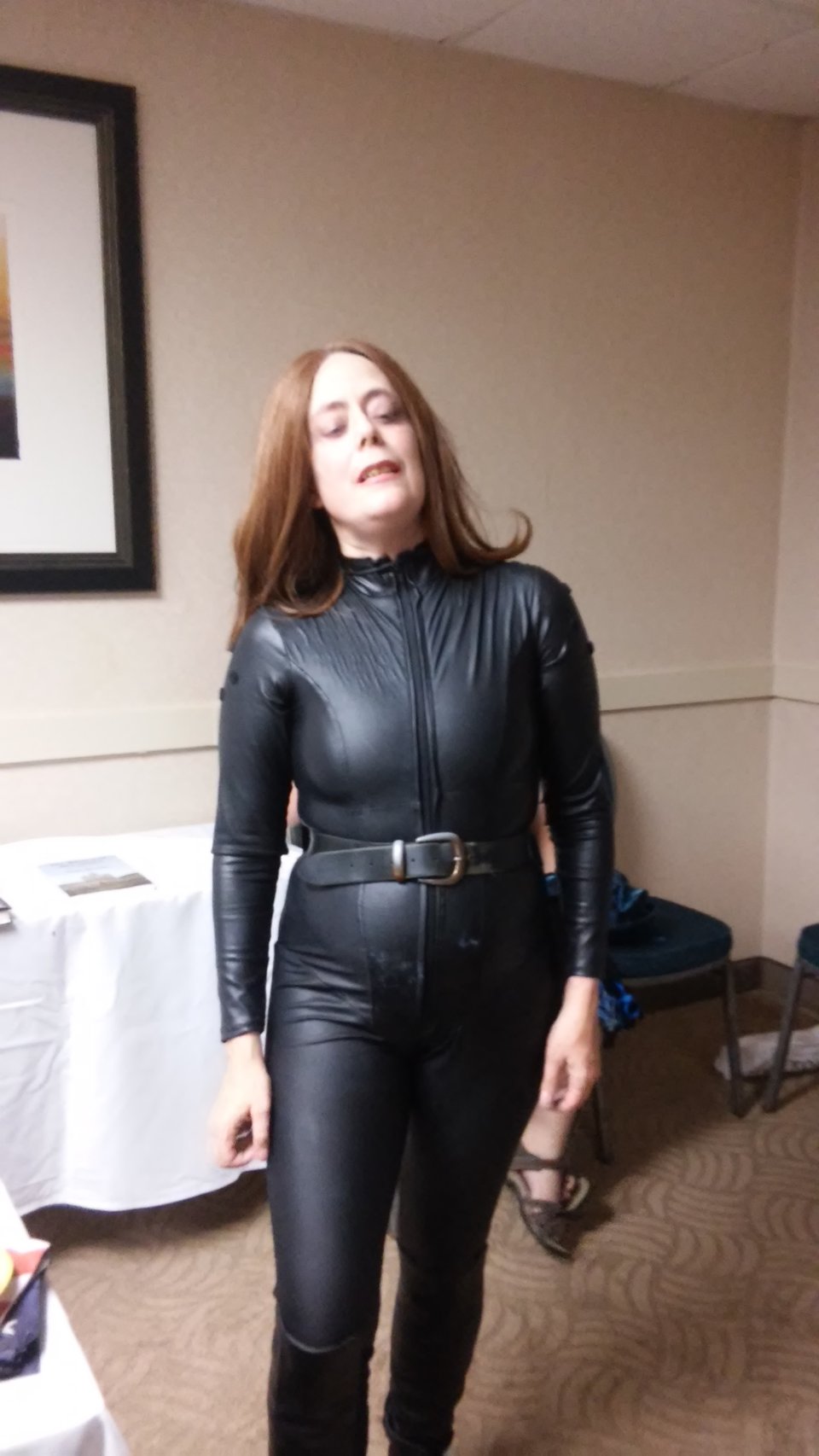 A woman wearing a black catsuit, belt, and gloves, with brown hair. Standing in a hotel conference room with white tables.