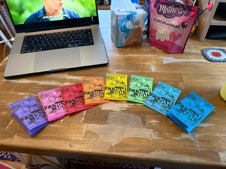 Picture of a rainbow spread of handmade, hand illustrated folded paper zines that say Family Night on the front. The zines are on a wooden table, with a laptop, box of kleenex, and bag of cookies in the background.
